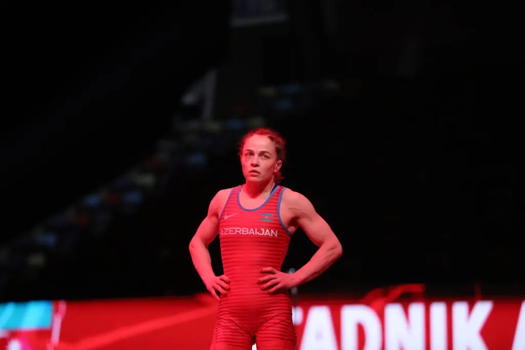 A women's wrestler stands in red light at the edge of the mat.